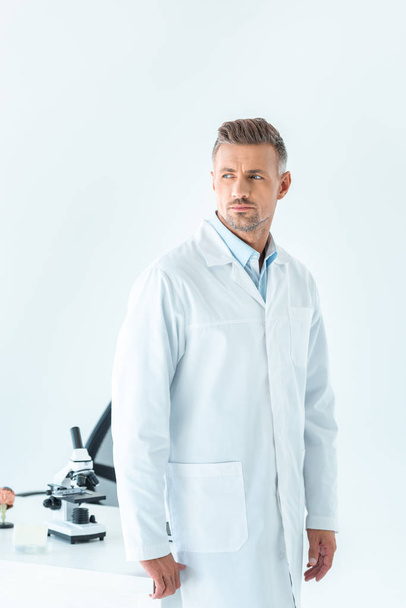 красивый ученый, стоящий рядом с столом с микроскопом и глядя в сторону изолированы на белом
 - Фото, изображение