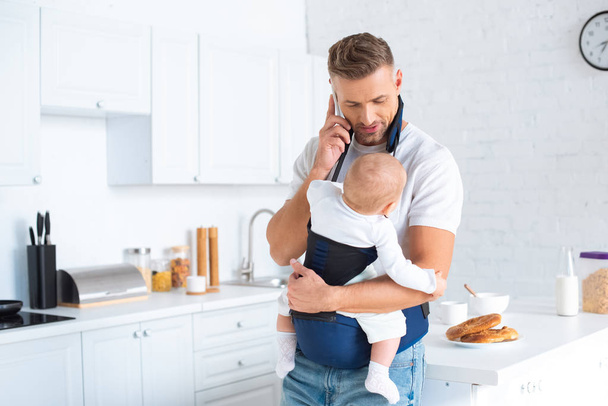 dad holding infant daughter in baby carrier and talking on smartphone in kitchen  - Valokuva, kuva
