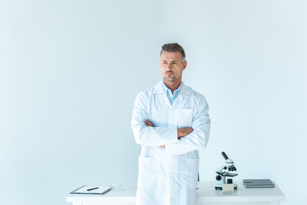 όμορφος επιστήμονας στέκεται με σταυρωμένα τα χέρια κοντά στο τραπέζι που απομονώνονται σε λευκό - Φωτογραφία, εικόνα