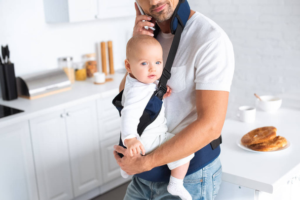 cropped view of father holding infant daughter in baby carrier and talking on smartphone  - Foto, Bild