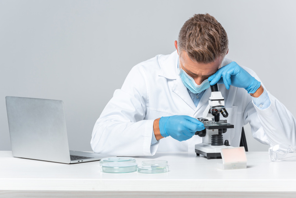 przystojny naukowiec w medycznych maski i Rękawice medyczne ogląda mikroskopu na białym tle - Zdjęcie, obraz