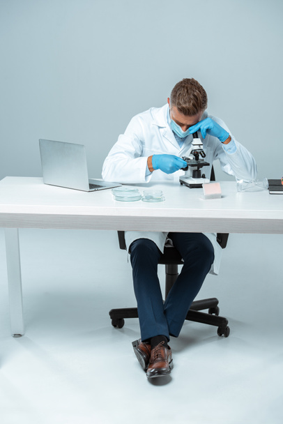 schöner Chemiker mit medizinischer Maske und medizinischen Handschuhen, der das Mikroskop isoliert auf weißem Grund betrachtet - Foto, Bild