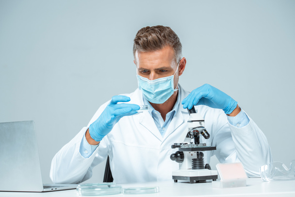 guapo científico en máscara médica y guantes médicos haciendo experimento aislado en blanco
 - Foto, Imagen