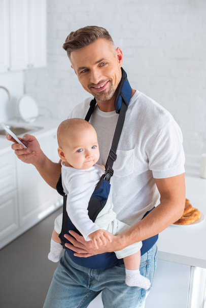 cheerful father using smartphone and holding infant daughter in baby carrier  - Photo, Image