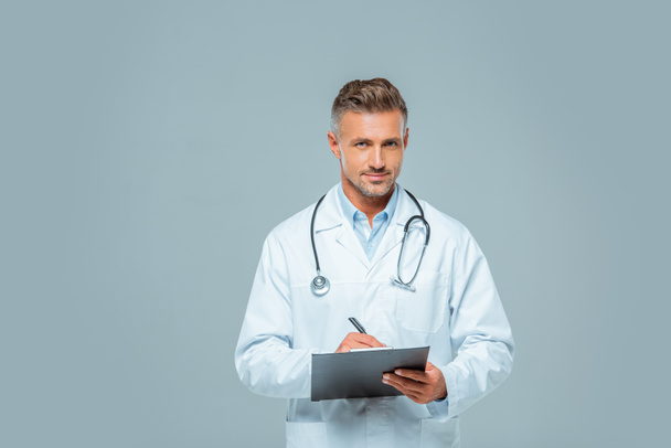 handsome doctor with stethoscope writing something to clipboard and looking at camera isolated on white - Photo, Image