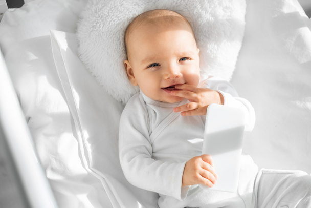 cheerful infant child holding smartphone in white baby crib  - Foto, Imagem