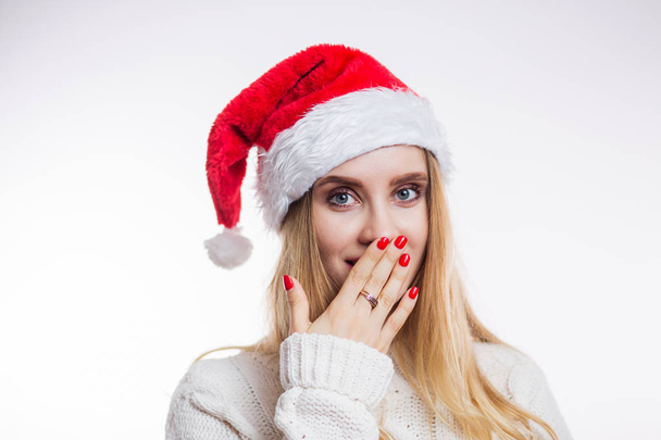 Portrait of surprised attractive young woman in red Santa hat and beige sweater on white background. A girl with a bright manicure closes her mouth with her hand and opens her eyes - Photo, image