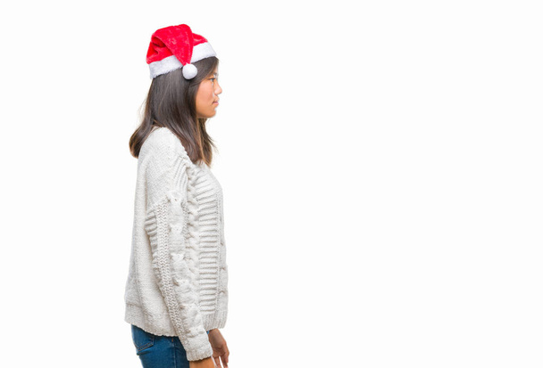 Giovane donna asiatica indossa cappello di Natale su sfondo isolato cercando di lato, posa profilo relax con viso naturale con sorriso fiducioso
. - Foto, immagini