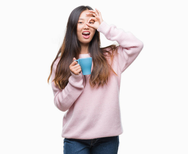 Young asian woman drinking coffee over isolated background with happy face smiling doing ok sign with hand on eye looking through fingers - Valokuva, kuva