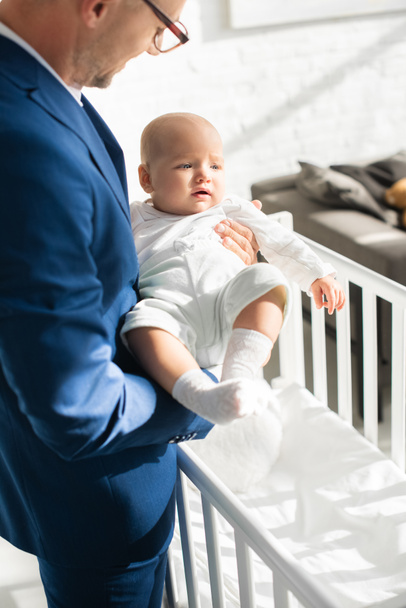 father in suit holding infant daughter near baby crib in room  - Fotoğraf, Görsel