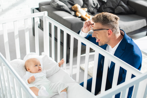 happy father in formal wear showing tongue to infant daughter in baby crib  - Fotoğraf, Görsel