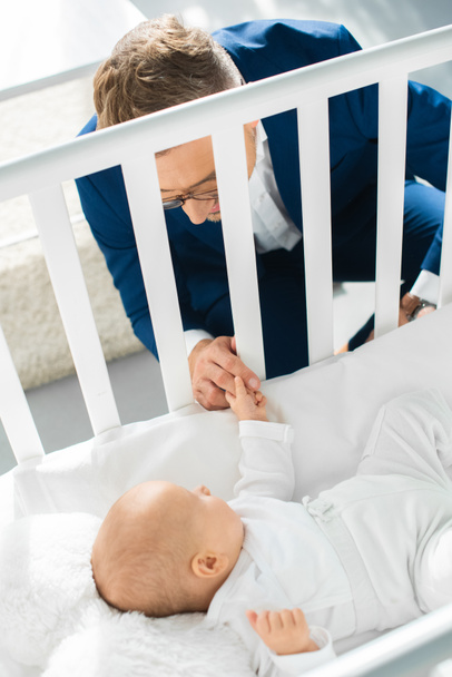 happy father in formal wear holding hand of infant daughter in baby crib  - Foto, Bild