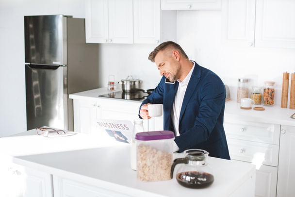 hombre de negocios sosteniendo taza con bebida mientras lee el periódico de negocios y habla en el teléfono inteligente en la cocina blanca
  - Foto, Imagen
