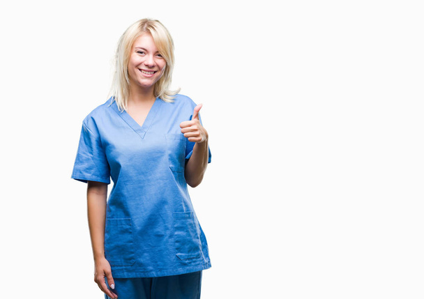 Young beautiful blonde nurse doctor woman over isolated background doing happy thumbs up gesture with hand. Approving expression looking at the camera with showing success. - Foto, Bild
