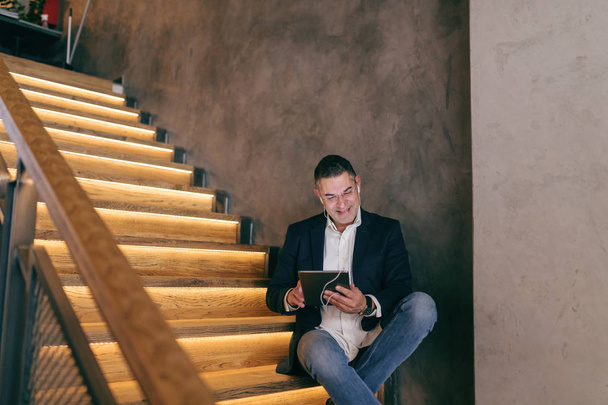 Smiling middle-aged Caucasian businessman dressed smart casual sitting on the stairs in cafe while using tablet for video call. Earphones in ears. - 写真・画像