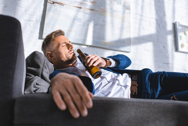 drunk businessman lying on sofa with closed eyes and drinking alcohol from bottle - Photo, Image