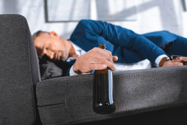 selective focus of bottle in hand of drunk businessman sleeping on sofa - Fotografie, Obrázek