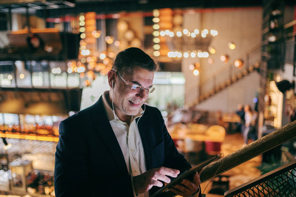 Primer plano de hombre de negocios sonriente de mediana edad vestido casual inteligente usando tableta para videollamada mientras está de pie en la cafetería. En los oídos auriculares
. - Foto, Imagen