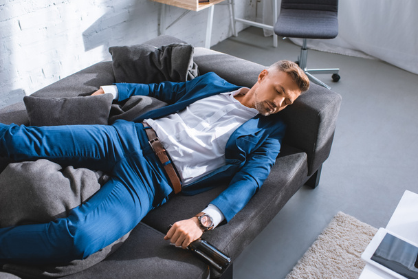 drunk businessman holding bottle and resting on sofa in living room  - Foto, Bild