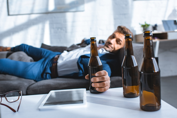 selective focus of bottle in hand of drunk businessman lying on sofa - Foto, afbeelding