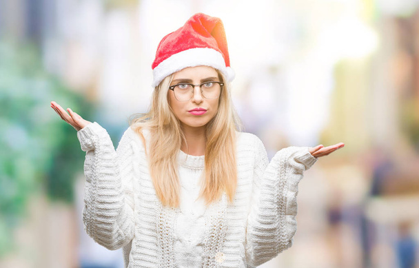 Fiatal gyönyörű szőke nő, fárasztó karácsonyi kalap alatt elszigetelt háttér tudatlan és zavaros kifejezés, a karok és a kezek. Kétség koncepció. - Fotó, kép