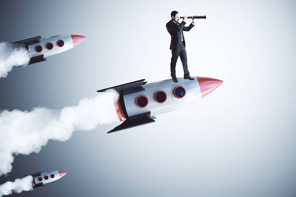 Businessman using telescope while standing on launching rocket. Gray background. Vision and startup concept  - Photo, Image