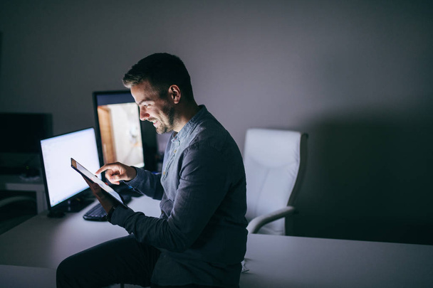 Χαμογελώντας Καυκάσιος γενειοφόρος επιχειρηματίας χρησιμοποιώντας tablet ενώ κάθεται στο γραφείο στο γραφείο αργά τη νύχτα. - Φωτογραφία, εικόνα
