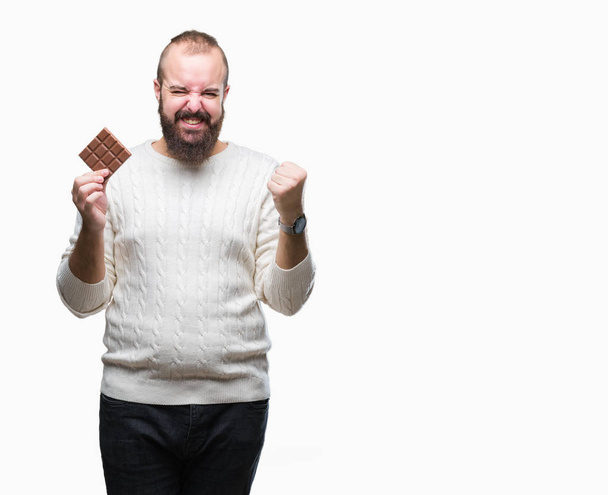 Joven hipster hombre comiendo barra de chocolate sobre fondo aislado gritando orgulloso y celebrando la victoria y el éxito muy emocionado, animando emoción
 - Foto, Imagen