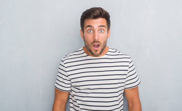 Handsome young man over grey grunge wall wearing navy t-shirt afraid and shocked with surprise expression, fear and excited face. - Photo, Image