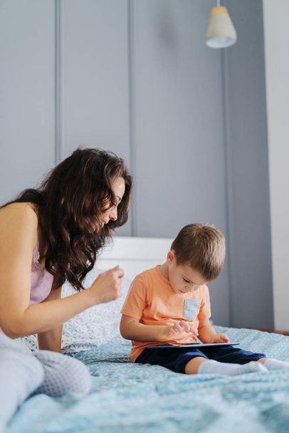 Little kid watching cartoons on tablet while sitting on the bed while his mother watching cartoons with him. Bedroom interior. - Φωτογραφία, εικόνα