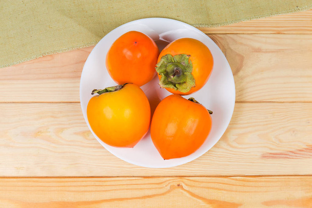 Vista superior de vários frutos de cáqui inteiros maduros frescos no prato branco na mesa rústica de madeira colorida clara
 - Foto, Imagem