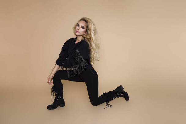 Beautiful, young female model in a autumn-winter clothing, military hoodie and overalls and boots on a beige background in the studio. Blonde in elegant biker, military style clothing - Foto, Bild