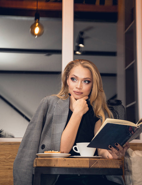 Beautiful young blonde woman with a cup of coffee and a book in a cafe - Fotografie, Obrázek