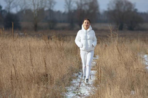 Preety smiling girl dressed all in white walks through the high dry grass field by snowy pathway at winter countryside - Photo, Image