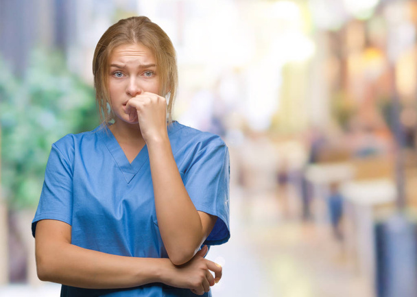 junge kaukasische Krankenschwester, die Chirurgenuniform über isoliertem Hintergrund trägt, sieht gestresst und nervös aus, die Hände auf mundgebissenen Nägeln. Angstproblem. - Foto, Bild