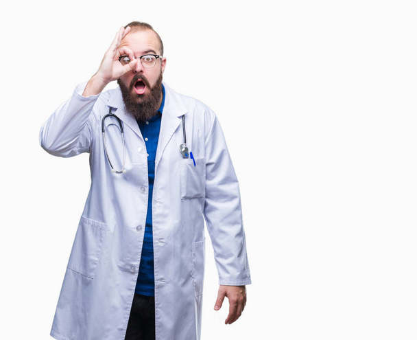 Молодий кавказький лікар чоловік в медичному білому пальто над ізольованим тлом робить нормальний жест шокованим здивованим обличчям, дивлячись крізь пальці. Невіруючий вираз
. - Фото, зображення