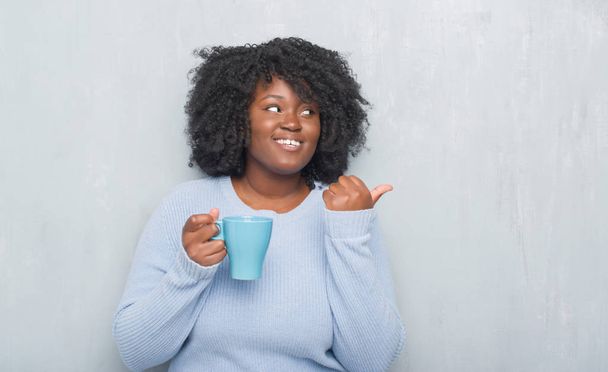 Jovem afro-americana sobre parede grunge cinza bebendo uma xícara de café apontando e mostrando com o polegar até o lado com o rosto feliz sorrindo
 - Foto, Imagem