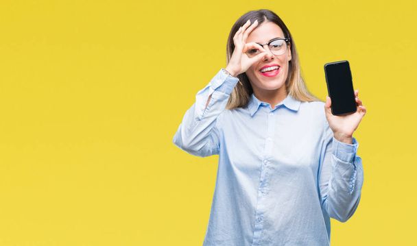 Молодая красивая деловая женщина показывает чистый экран смартфона на изолированном фоне с счастливым лицом улыбается делает хорошо знак с рукой на глазу глядя сквозь пальцы
 - Фото, изображение