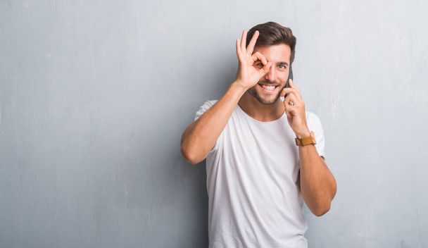 Handsome young man over grey grunge wall speaking on the phone with happy face smiling doing ok sign with hand on eye looking through fingers - Foto, Bild