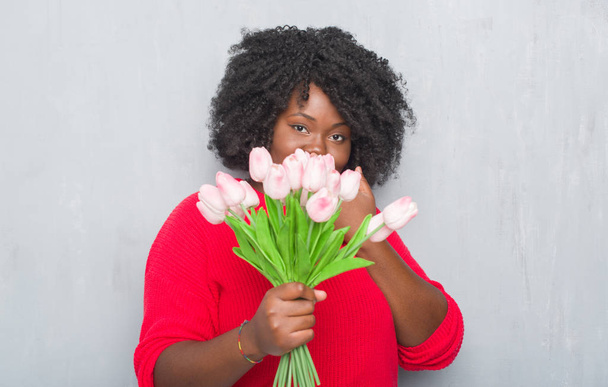 Joven mujer afroamericana sobre pared grunge gris sosteniendo flores rosadas ramillete cubrir la boca con la mano conmocionado con vergüenza por error, expresión de miedo, asustado en silencio, concepto secreto
 - Foto, imagen