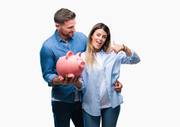 Young couple in love holding piggy bank over isolated background with surprise face pointing finger to himself - Fotoğraf, Görsel