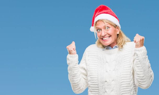 Middle age blonde woman wearing christmas hat over isolated background very happy and excited doing winner gesture with arms raised, smiling and screaming for success. Celebration concept. - Photo, Image