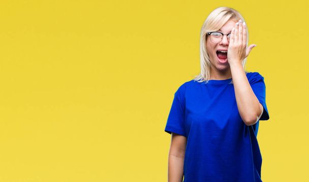 Young beautiful blonde woman wearing glasses over isolated background covering one eye with hand with confident smile on face and surprise emotion. - Photo, Image