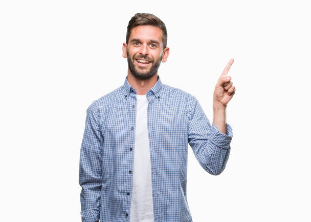 Молодий красивий чоловік у білій футболці над ізольованим тлом з великою посмішкою на обличчі, вказуючи рукою і пальцем в сторону, дивлячись на камеру
. - Фото, зображення