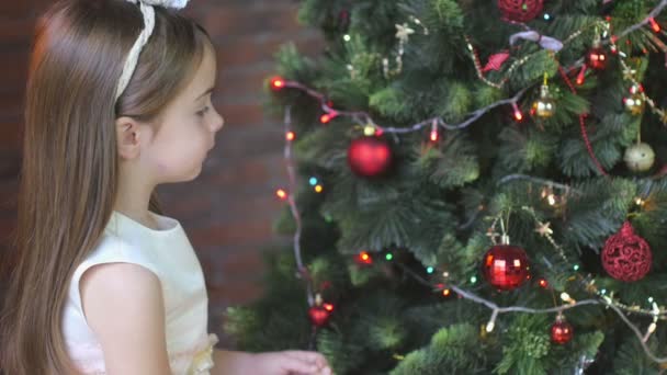 beautiful little girl in a festive dress decorates a Christmas tree with balls - Filmagem, Vídeo