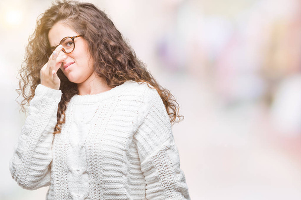 Beautiful brunette curly hair young girl wearing winter sweater over isolated background smelling something stinky and disgusting, intolerable smell, holding breath with fingers on nose. Bad smells concept. - Photo, Image