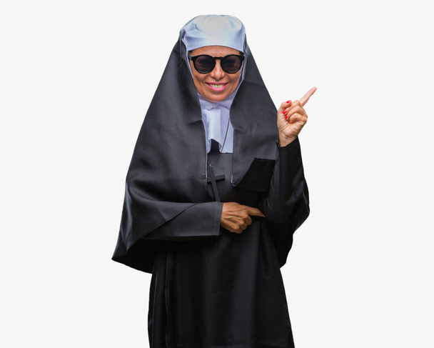 Femme religieuse catholique âgée du Moyen Âge portant des lunettes de soleil sur fond isolé avec un grand sourire sur le visage, pointant la main et le doigt vers le côté en regardant la caméra
. - Photo, image