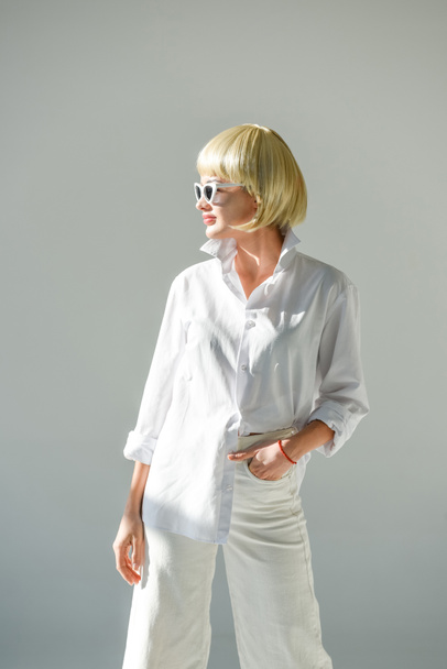 attractive blonde woman in sunglasses and stylish white outfit looking away on white - 写真・画像