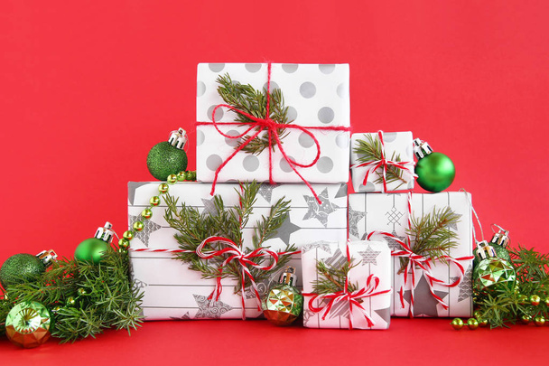 Christmas gift boxes wrapped of white-gray paper with red-white ribbons on a red background, decorated of fir branches and shiny green Christmas decorations. Christmas and New Year concept with copy space. - Photo, Image