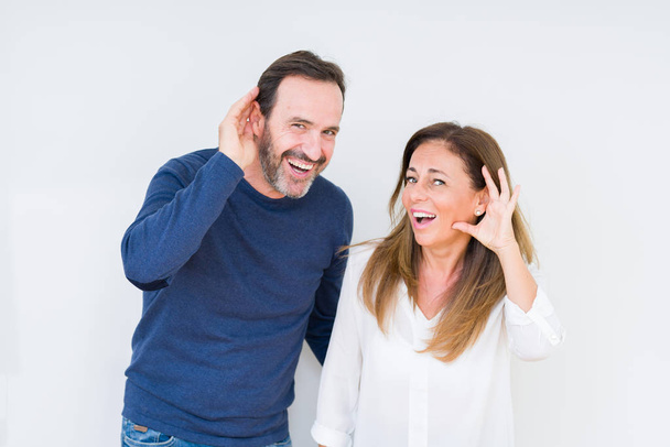 Beautiful middle age couple in love over isolated background smiling with hand over ear listening an hearing to rumor or gossip. Deafness concept. - Photo, Image